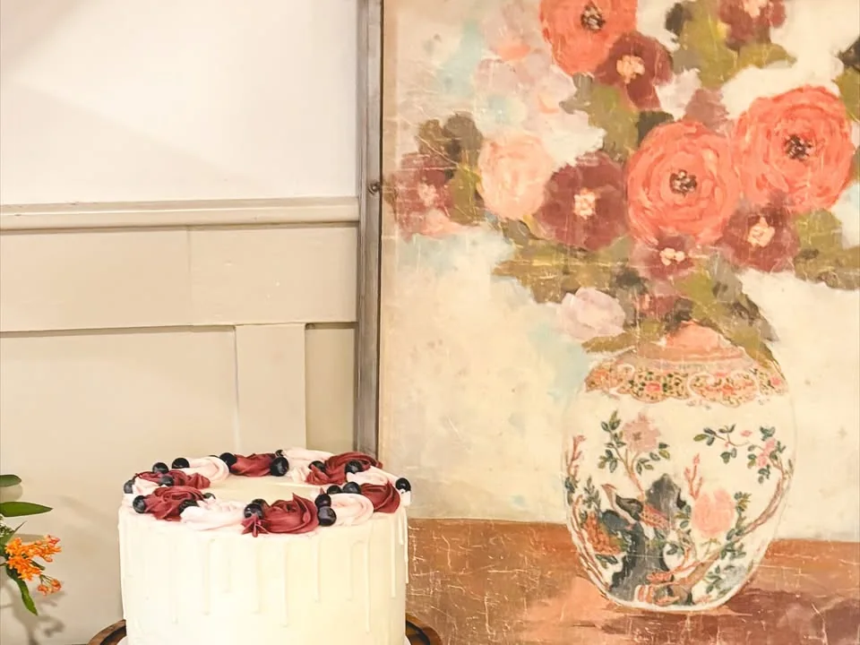 Single-tier white cake displayed on a pedestal beside floral artwork.
